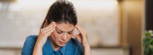 A imagem mostra uma mulher com expressão de cansaço ou estresse. Ela está com os olhos fechados e as sobrancelhas franzidas, pressionando as têmporas com as mãos, sugerindo que está sentindo dor de cabeça ou sobrecarga mental. A mulher veste uma blusa azul e está em um ambiente interno com iluminação suave ao fundo. A cena transmite uma sensação de exaustão ou tensão emocional