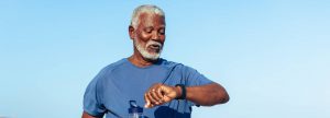 A imagem mostra um homem idoso de barba e cabelo grisalhos, vestido com uma camiseta esportiva azul. Ele segura uma garrafa de água na mão esquerda e olha para um relógio ou pulseira inteligente em seu pulso direito. O céu ao fundo está limpo e azul, sugerindo que ele está ao ar livre em um dia ensolarado. O homem parece estar praticando atividades físicas ou caminhando, transmitindo uma sensação de saúde, bem-estar e vitalidade.