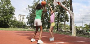A imagem mostra duas pessoas jogando basquete em uma quadra ao ar livre. A pessoa à esquerda está vestindo uma camiseta verde e shorts claros, segurando a bola de basquete e se preparando para arremessar. A pessoa à direita, vestindo uma camiseta rosa e shorts amarelos, está tentando bloquear o arremesso com o braço levantado. O cenário de fundo inclui árvores altas, alguns prédios residenciais e um céu parcialmente nublado. A quadra tem piso avermelhado e a cesta de basquete está montada em uma estrutura branca.