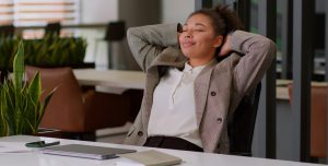 A pessoa veste um blazer xadrez e uma camisa branca, transmitindo um visual profissional. À sua frente, sobre a mesa, há um notebook fechado, um smartphone e alguns papéis. O ambiente parece ser um escritório moderno, com plantas e mobiliário elegante ao fund
